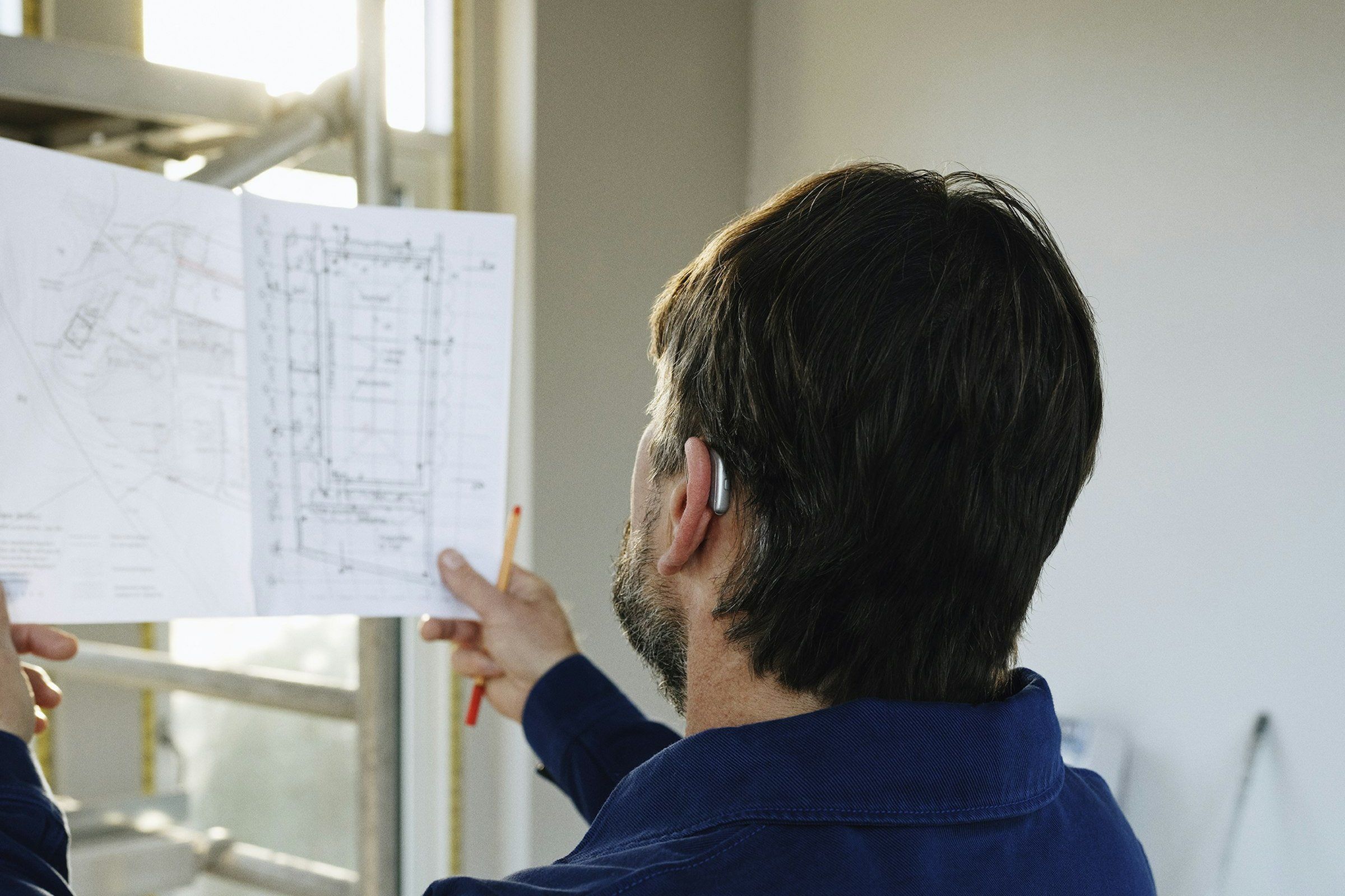 A builder holds up a set of plans with a pencil in hand, inside a plastered house still under construction