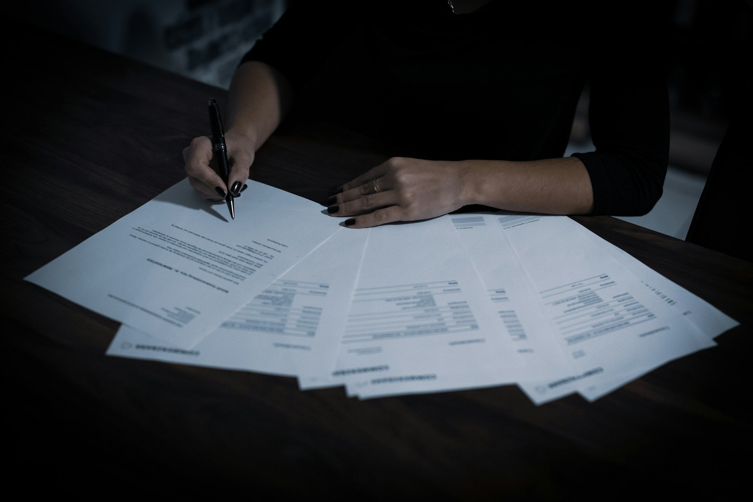 A person signing one of several printed documents spread across a dark table
